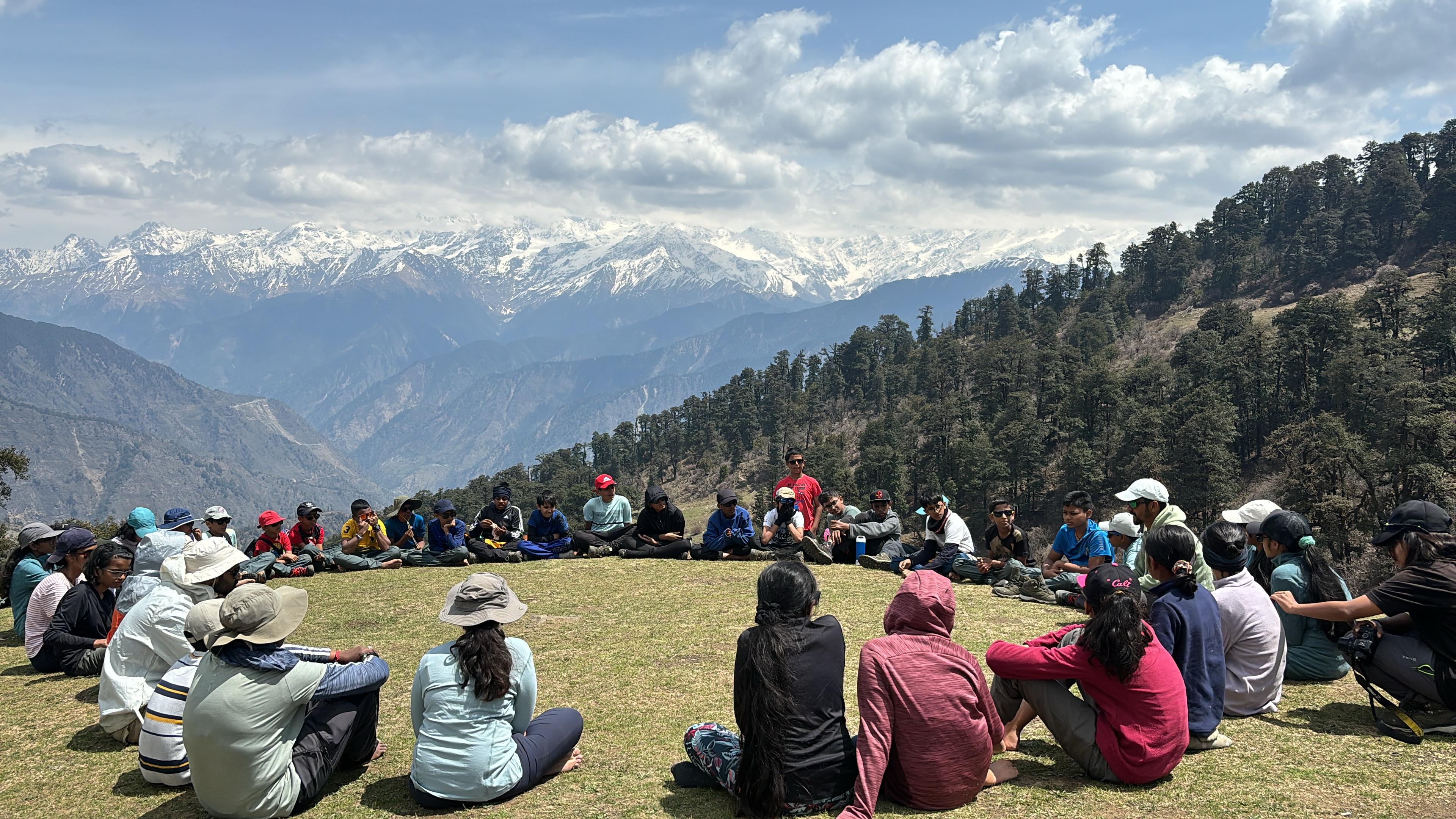 The trek group sitting in a circle on a meadow with snow-capped Himalayan peaks behind